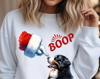 Op de afbeelding: Wit sweatshirt met een kerstmanhand die de neus van een hond aantikt, met het woord "BOOP" in het rood. De hond is zwart, bruin en wit. Het sweatshirt wordt gedragen met grijze shorts.