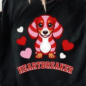 May include: Black t-shirt featuring a cartoon dachshund with pink and red stripes, surrounded by hearts. The word "HEARTBREAKER" is printed in red and white. A white mug with a red heart is held in the upper left.