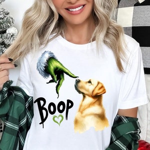 May include: White t-shirt with a green Grinch hand reaching out to a golden retriever dog. The text "Boop" is printed on the shirt with a green heart.
