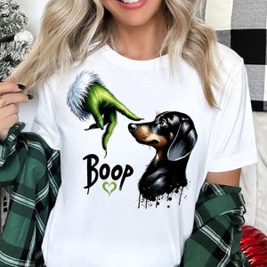 Puede incluir: Camiseta blanca con una mano verde del Grinch que se extiende hacia un dachshund negro y marrón. El texto "Boop" está impreso en la camiseta con un corazón verde.