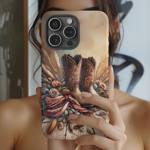 May include: A phone case featuring a rustic design with brown cowboy boots, flowers, and wheat stalks. The case has a wooden plank background and a soft, neutral color palette. The design evokes a country or western theme.