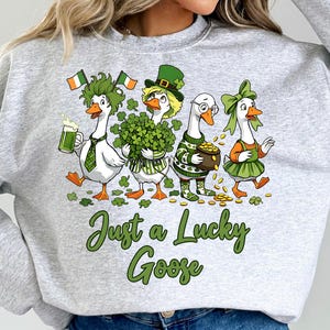 May include: A grey sweatshirt with a St Patrick's Day design. Four cartoon geese are depicted, dressed in green and gold, holding items like beer, shamrocks, and a pot of gold. The text "Just a Lucky Goose" is written in green.