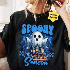 Könnte beinhalten: Schwarzes T-Shirt mit einem Halloween-Motiv. Das Design zeigt das Wort "Spooky" in Blau, einen lächelnden Geist, ein Spukhaus und das Wort "Season" in Blau. Blitze und kleine Geister sind ebenfalls abgebildet.