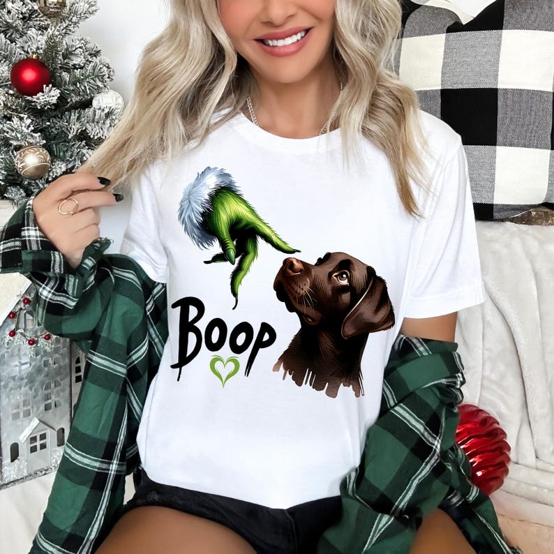 以下が含まれることがあります： 白いTシャツ。緑色の手と白い毛皮、茶色の犬、「Boop」の文字と緑色のハートが描かれています。カジュアルなスタイルのTシャツです。