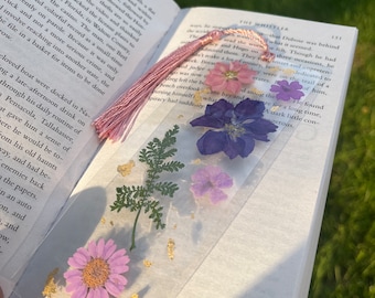 Könnte beinhalten: Ein transparentes Lesezeichen mit gepressten Blumen und goldenen Blatt-Akzenten. Die Blumen sind lila, rosa und weiß. Das Lesezeichen hat einen rosa Quasten.