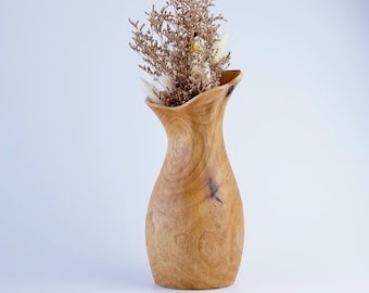 May include: A light brown wooden vase with a smooth, curved silhouette and visible natural wood grain patterns. The vase contains a cluster of dried, light brown and white flowers with small, delicate stems and tiny petals extending upward.