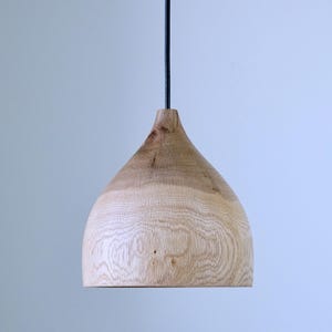 Peut inclure: Un luminaire suspendu en bois avec un grain de bois naturel. La lampe a une forme de goutte d'eau et est suspendue par un cordon noir. Le luminaire mesure environ 25 cm de haut et 20 cm de large.