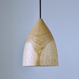 Könnte beinhalten: Eine Holz-Pendelleuchte mit konischem Schirm. Der Schirm besteht aus hellem Holz mit sichtbaren Maserungsmustern. Ein schwarzes Kabel hängt die Leuchte von oben auf. Die Leuchte befindet sich vor einem hellblauen Hintergrund.