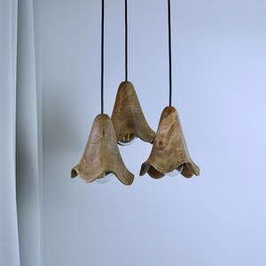Peut inclure: Trois lampes suspendues en bois avec une finition naturelle veinée. Chaque lampe a un abat-jour en forme de cloche avec un bord évasé, suspendu par un cordon noir. Les lampes sont suspendues sur un fond blanc.