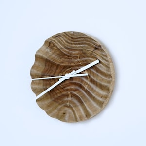 Peut inclure: Une horloge ronde en bois avec un motif de grain naturel. L'horloge est de couleur marron clair avec des aiguilles blanches. Le cadran est une coupe transversale d'un tronc d'arbre. L'horloge est fixée sur un mur blanc.