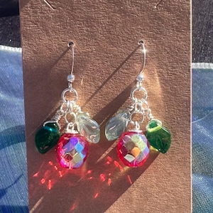 Puede incluir: Un par de pendientes colgantes con ganchos plateados. Cada pendiente presenta una cuenta rosa iridiscente facetada, dos cuentas transparentes en forma de hoja y una cuenta verde en forma de lágrima. Los pendientes se exhiben en una tarjeta marrón.