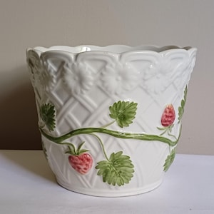 May include: White ceramic plant pot with a woven basket pattern and a scalloped edge. It features green vines, leaves, and red strawberries. A decorative item for the home or garden.