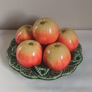 May include: A ceramic fruit bowl featuring five apples with a gradient of yellow to red hues. The apples rest on a green, leaf-shaped ceramic dish. The arrangement is a decorative piece.