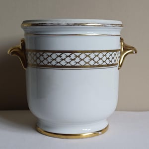 May include: White ceramic ice bucket with gold trim and handles. The bucket has a gold geometric pattern around the middle and gold accents on the rim and base. The handles are ornate and gold. A classic piece for serving beverages.