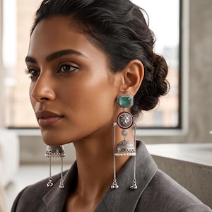 May include: A pair of long silver earrings featuring a rectangular green stone at the top, a circular red accent with a central lion head motif, and a bell-shaped jhumka base with small white beads and dangling silver chains at the bottom.