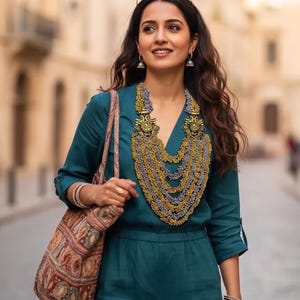 May include: A teal jumpsuit with a multi-layered gold and silver beaded statement necklace, small silver jhumka earrings, and a patterned fabric tote bag. The outfit is accessorized with stacked metal bangles, a gold ring, and sunglasses on the head.
