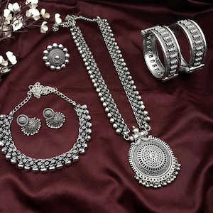 Op de afbeelding: Een zilverkleurige sieradenset met een ketting, oorbellen, ring en twee armbanden. De ketting heeft een grote, ronde hanger met ingewikkelde details. De set wordt gepresenteerd op een bordeauxrode satijnen stof.