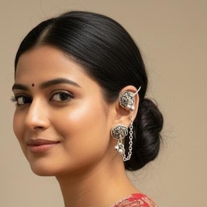 May include: A woman wearing a red and gold embroidered top and silver ear cuff earrings. The earrings feature a floral design and a chain connecting to a stud earring. The woman has dark hair and is looking at the camera.