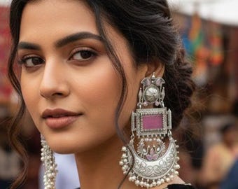 May include: A woman wearing large, ornate silver earrings with a rectangular pink stone and a crescent moon design. The earrings have intricate detailing and small pearl accents. The woman is wearing a black top.