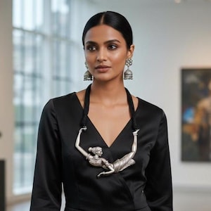 Può includere: Una donna indossa una tuta nera e gioielli d'argento, tra cui orecchini e una collana. La collana presenta una figura d'argento. Lo sfondo è una galleria d'arte con dipinti e sculture.