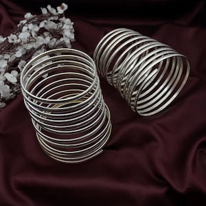 Może przedstawiać: Dwa zestawy srebrnych bransoletek ułożonych jedna na drugiej na tle burgundowego materiału. Bransoletki są cienkie i mają gładkie, błyszczące wykończenie.