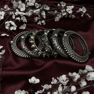 May include: A set of five silver-toned bangles with intricate floral and geometric designs. The bangles are stacked on a burgundy fabric background.