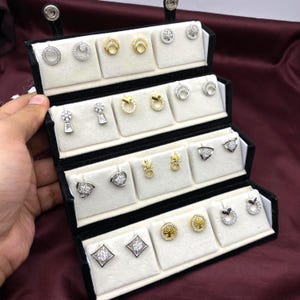 Puede incluir: Un joyero negro abierto que muestra varios pares de pendientes. Los pendientes son plateados y dorados, con diseños que incluyen círculos, flores y formas geométricas. Los pendientes se exhiben sobre almohadillas individuales de terciopelo blanco.