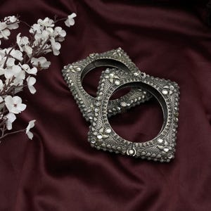 May include: Two silver square-shaped bangles with intricate designs and small round stones. The bangles are on a dark red fabric background.