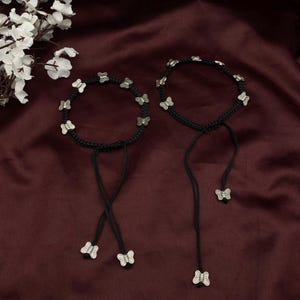 Op de afbeelding: Twee zwarte touwen enkelbandjes met zilveren vlinder bedels. De enkelbandjes zijn gebonden met een strik aan de enkel.
