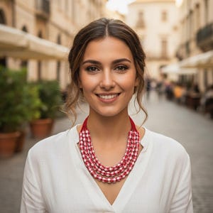 Könnte beinhalten: Mehrreihige Halskette mit roten und weißen Perlen. Die Halskette ist mehrschichtig und hat ein auffälliges Design. Die Frau trägt ein weißes Hemd. Der Hintergrund ist eine verschwommene Straßenszene.