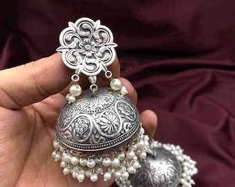 May include: Silver-toned jhumka earrings with intricate floral and leaf patterns. The earrings feature a bell-shaped design, adorned with small white pearls. The top portion has a floral design, and the bottom is embellished with dangling pearls.