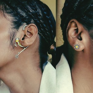 May include: A close-up of a person's ear with two earrings. The left ear has a silver ear cuff with a yellow and black bird design. The right ear has a silver flower-shaped stud with a yellow center.