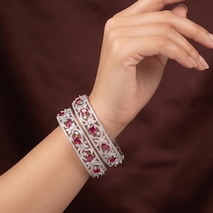 May include: Two silver-toned metal bangles featuring an intricate openwork floral pattern. Each bangle is set with multiple pear-shaped red stones and small clear accent stones embedded throughout the filigree design, creating a detailed look.