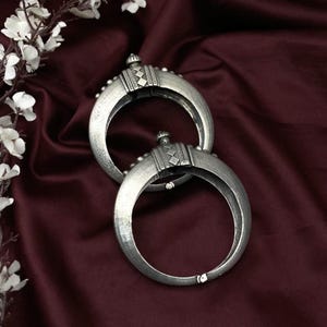 May include: Two silver crescent bangles with detailed geometric patterns. The bangles are displayed on a deep burgundy satin background, with white flowers in the top left corner. The jewellery has a vintage or antique aesthetic.
