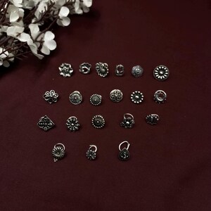 Pu&ograve; includere: Una collezione di 18 anelli per il naso in argento con vari design, tra cui fiori, forme geometriche e un pavone. Gli anelli sono disposti in file su uno sfondo rosso scuro.