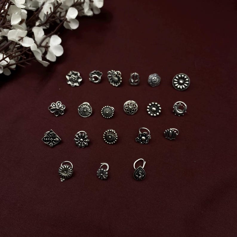 Pu&ograve; includere: Una collezione di 18 anelli per il naso in argento con vari design, tra cui fiori, forme geometriche e un pavone. Gli anelli sono disposti in file su uno sfondo rosso scuro.