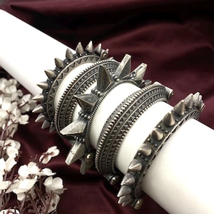 Może przedstawiać: Kolekcja srebrnych bransoletek o estetyce punk rock. Bransoletki mają różne wzory kolców i ćwieków, ze skomplikowanymi wytrawionymi wzorami. Biżuteria jest prezentowana na białym cylindrycznym kształcie na bordowym tle.