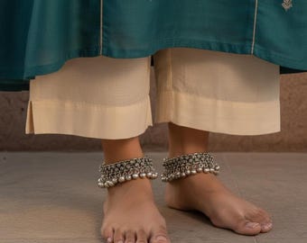 May include: A pair of silver anklets with dangling bells adorn the ankles. The anklets feature intricate detailing and are worn with cream-colored cropped pants. The outfit is paired with a teal top, creating a contrast of colors.
