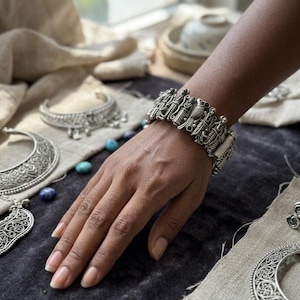 May include: An intricately designed silver bracelet, adorned with bird and leaf patterns. The bracelet is worn on a hand, with other silver jewellery pieces in the background. The jewellery is displayed on a dark surface with fabric.
