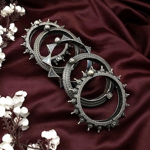Puede incluir: Cuatro pulseras plateadas con intrincados diseños y tachuelas decorativas. Las pulseras están dispuestas sobre una tela de satén burdeos oscuro, con flores blancas en la esquina. Las pulseras tienen una estética vintage o antigua.