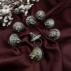 Könnte beinhalten: Eine Sammlung verzierter Silberringe mit farbigen Edelsteinakzenten. Die Ringe weisen aufwendige Designs auf und sind auf einem tief burgunderfarbenen Stoff platziert. Einige Ringe haben eine Kuppelform, andere ein traditionelleres Ringdesign.