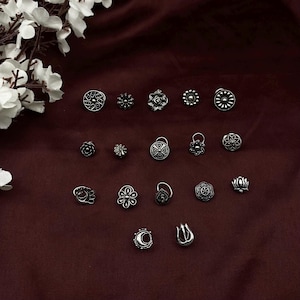 Pu&ograve; includere: Una collezione di 18 anelli per il naso in argento con vari disegni intricati, tra cui motivi floreali, geometrici e astratti. Gli anelli sono disposti su uno sfondo di tessuto marrone scuro.