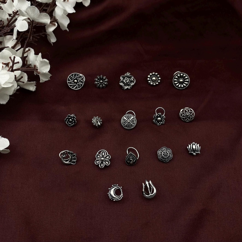 Pu&ograve; includere: Una collezione di 18 anelli per il naso in argento con vari disegni intricati, tra cui motivi floreali, geometrici e astratti. Gli anelli sono disposti su uno sfondo di tessuto marrone scuro.