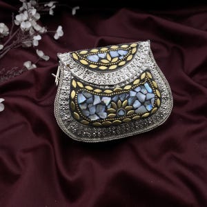 May include: Silver-toned metal clutch with intricate detailing. The purse features a mosaic of iridescent blue and white stones, framed by gold-toned accents. The design includes floral and leaf patterns, creating a decorative and elegant accessory.