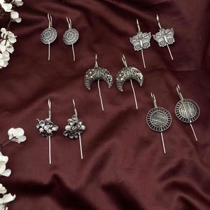 May include: An assortment of silver-toned earrings with various designs, including circular, butterfly, crescent moon, and floral shapes. The earrings are displayed on a burgundy fabric background, showcasing their intricate details and craftsmanship.
