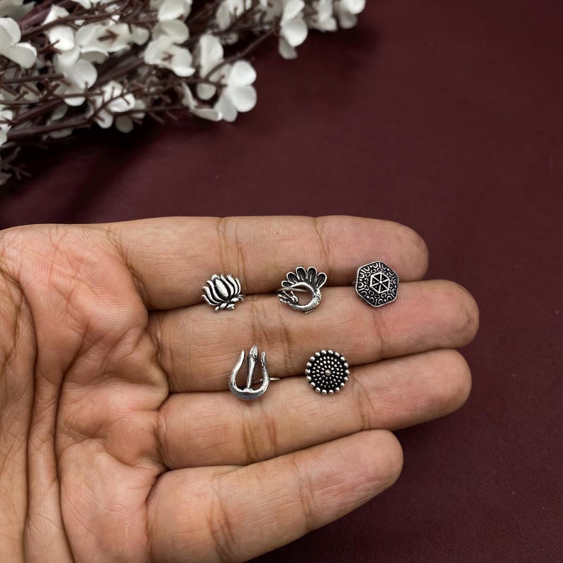 Pu&ograve; includere: Set di cinque anelli per il naso in argento con diversi design, tra cui un fiore di loto, un pavone, un motivo geometrico, un tridente e un design rotondo.