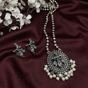 Puede incluir: Un collar de plata con un colgante redondo que presenta una imagen detallada de una figura tocando una flauta. El colgante está rodeado por un borde decorativo y cuelga de una cadena con acentos de perlas blancas. El collar viene con un par de aretes a juego que presentan el mismo diseño de figura.