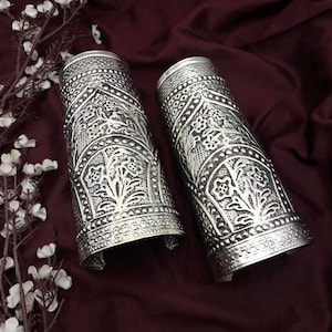 May include: Two silver-colored arm cuffs with intricate floral and geometric patterns. The cuffs are cylindrical with an open design, showcasing detailed embossed designs. The background is a deep burgundy fabric, with white flowers in the upper left corner.