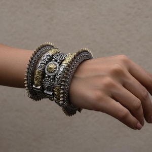 Könnte beinhalten: Eine Sammlung verzierter Armreifen in Silber- und Goldtönen. Die Armreifen weisen komplizierte Muster, darunter florale und geometrische Designs, sowie kleine, erhabene Details auf. Die Armbänder sind an einem Handgelenk gestapelt und zeigen ihre unterschiedlichen Texturen und Stile.