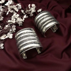 Peut inclure: Deux bracelets manchette argentés avec des motifs en relief complexes. Chaque bracelet comporte plusieurs bandes avec des motifs détaillés, notamment floraux et pointillés. Les bracelets sont présentés sur un tissu bordeaux foncé, avec des fleurs blanches en arrière-plan.